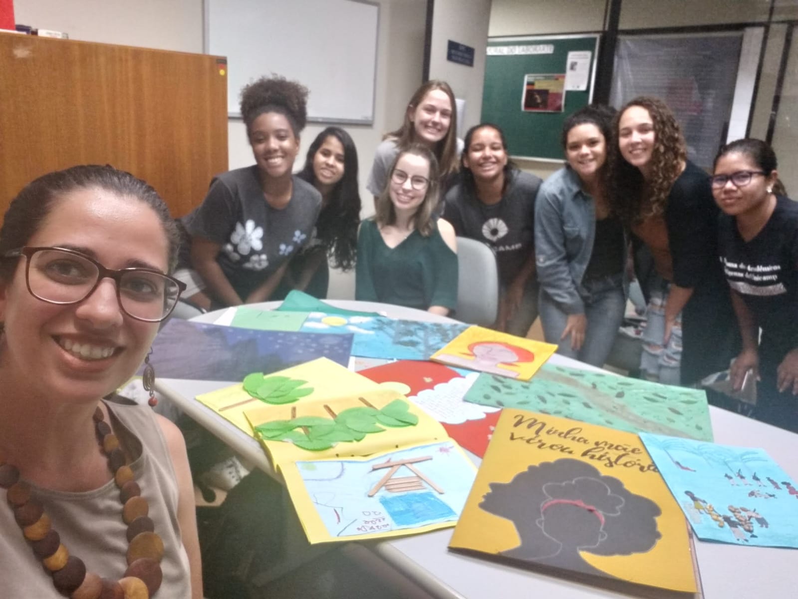 Fotografia horizontal de um pequeno grupo de estudantes de graduação em uma sala de aula ao redor de uma mesa, na qual espalham-se cartazes coloridos. Afrente, a Profa. Gabriela Tebet registra a foto como selfie. Ela é uma mulher parda, cabelos cacheados pretos amarrados para trás, óculos de grau, blusa bege, colar colorido. Estudantes são todas mulheres jovens, algumas negras, brancas e uma indígena, com cabelos e roupas diversos.