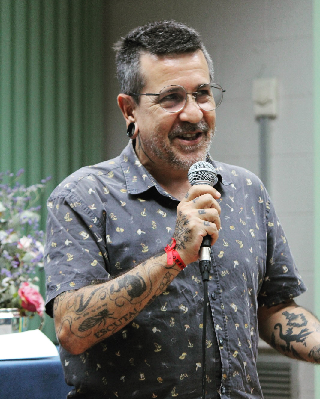 Retrato vertical do Prof. Andre Luiz Correia Gonçalves de Oliveira. Ele é um homem pardo, baixo, caelos curstos grisalhos, olhos amendoados, usa óculos de grau de armação metálica, alargador na orelha direita, possui tatuagens nos braços, veste camisa cinza com estampas brancas. Gesticula ao falar em um microfone. Ao fundo, auditório com parede branca e cortinas venezianas verdes.