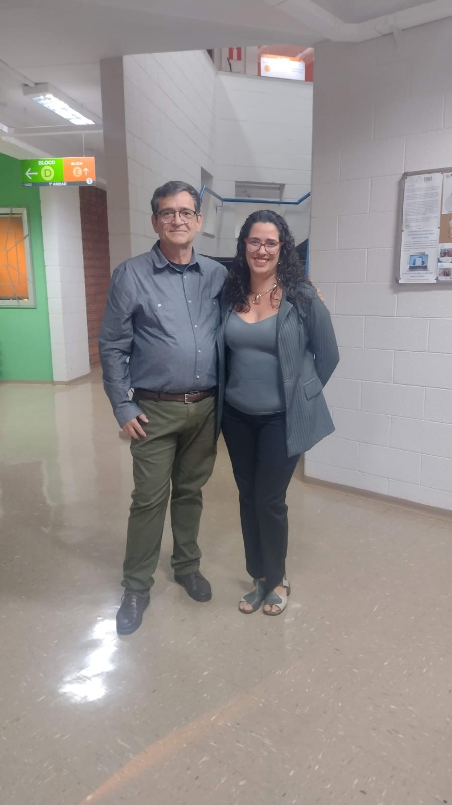 Fotografia frontal dos coordenadores do Curso de Pedagogia da Unicamp. Prof. Carlos Miranda é um homem branco de estatura média, rosto redondo, cabelos curtos acinzentados, usa óculos de grau, camisa cinza de mangas compridas, calça verde oliva, sapatos escuros. Profa. Gabriela Tebet é uma mulher parda, com cabelos longos escuros cacheados abaixo dos ombros, usa óculos de grau de armação vermelha, blusa cinza, paletó cinza, calça preta, sandálias de couro cinza. Os dois estão em pé em meio a um corredor.