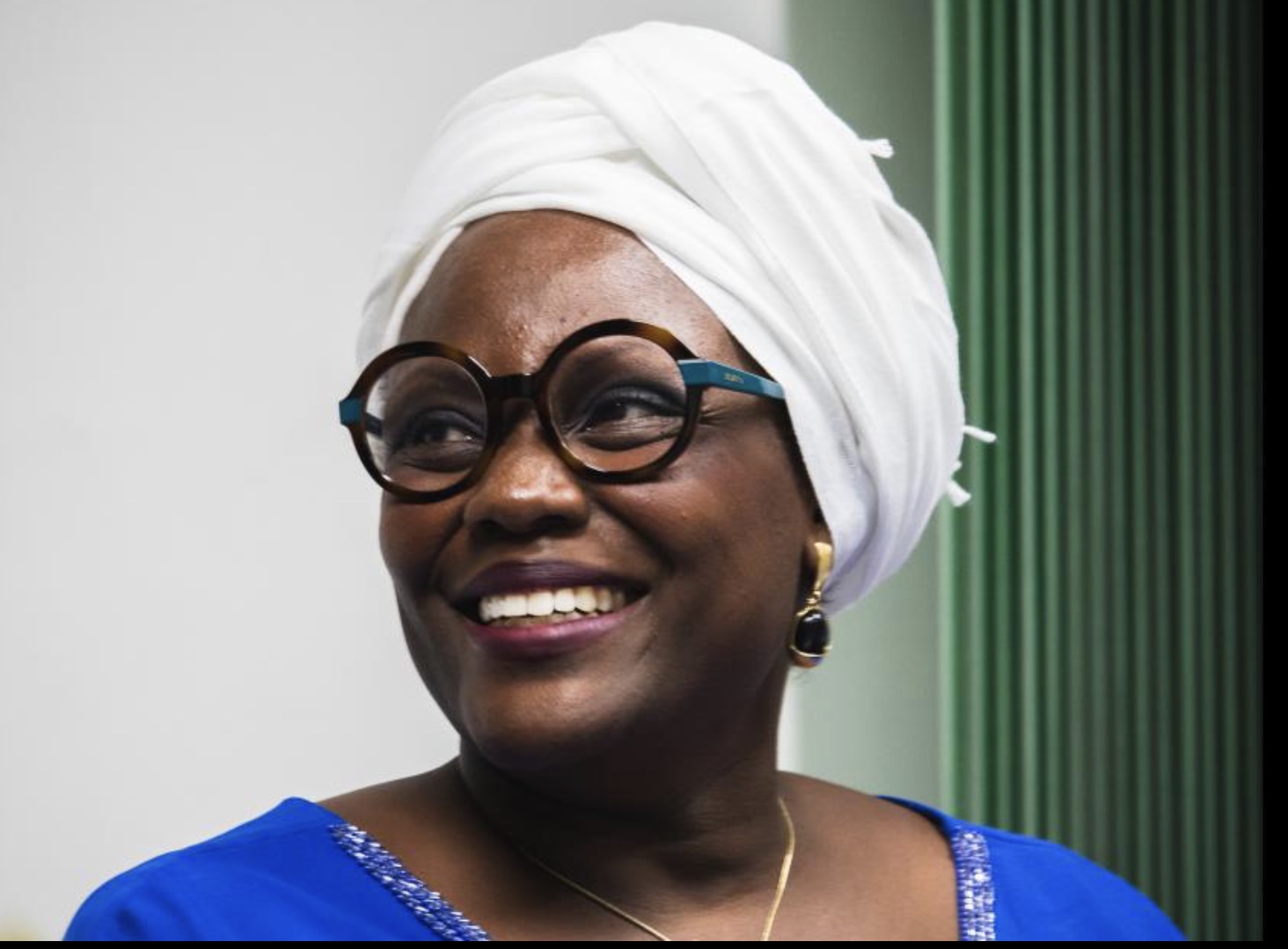 Retrato horizontal da Professora Debora Jeffrey, uma mulher negra retinta sorridente, com turbante branco sobre os cabelos, óculos de grau de aro redondo preto e vestido azul vivo. Ao fundo parede branca com cortinas venezianas verdes.