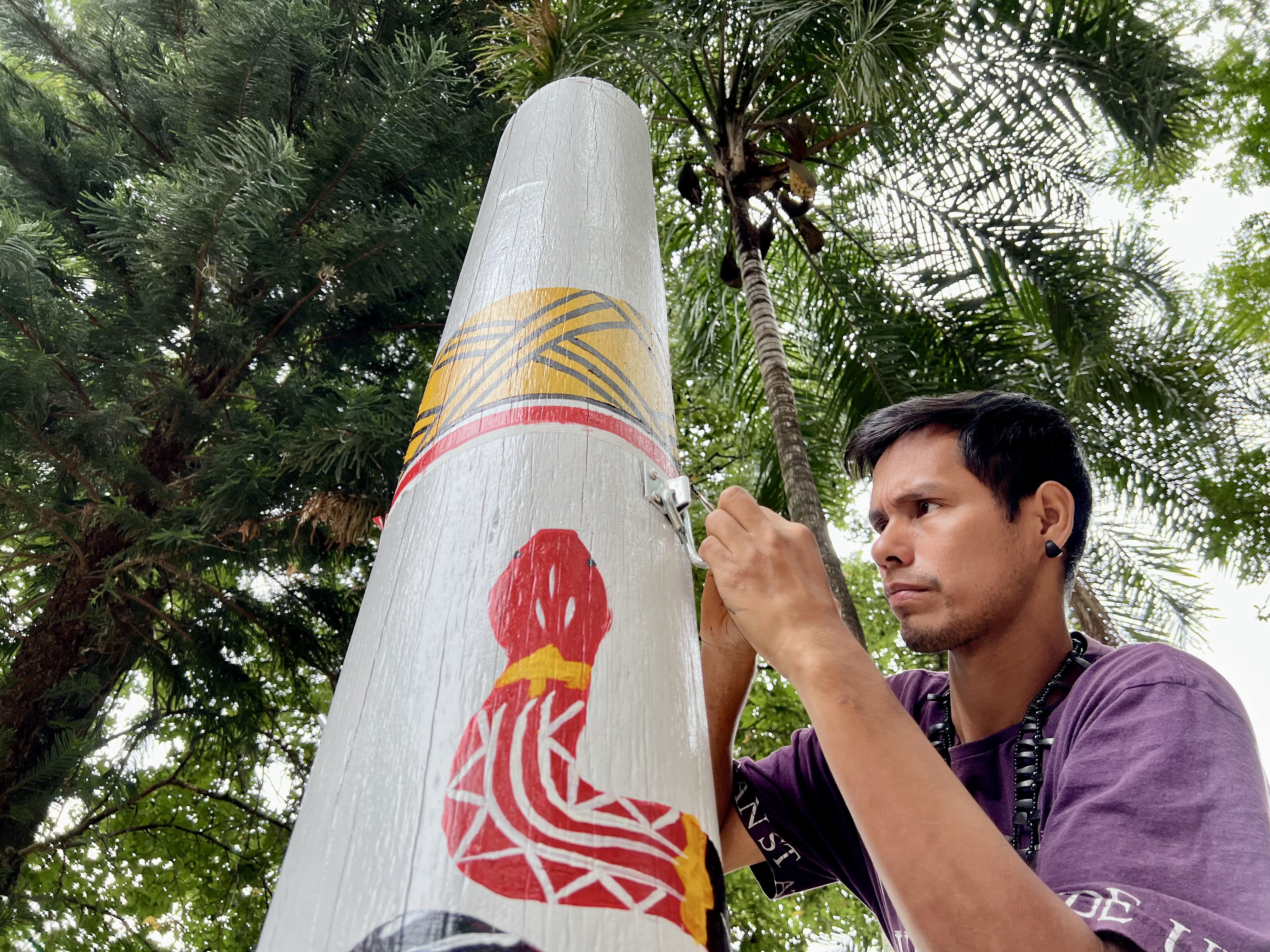 Fotografia de um rapaz indígena pintando com tinta um mastro branco com grafismos coloridos de representação de cobra vermelha. Ao redor, copa de árvores.