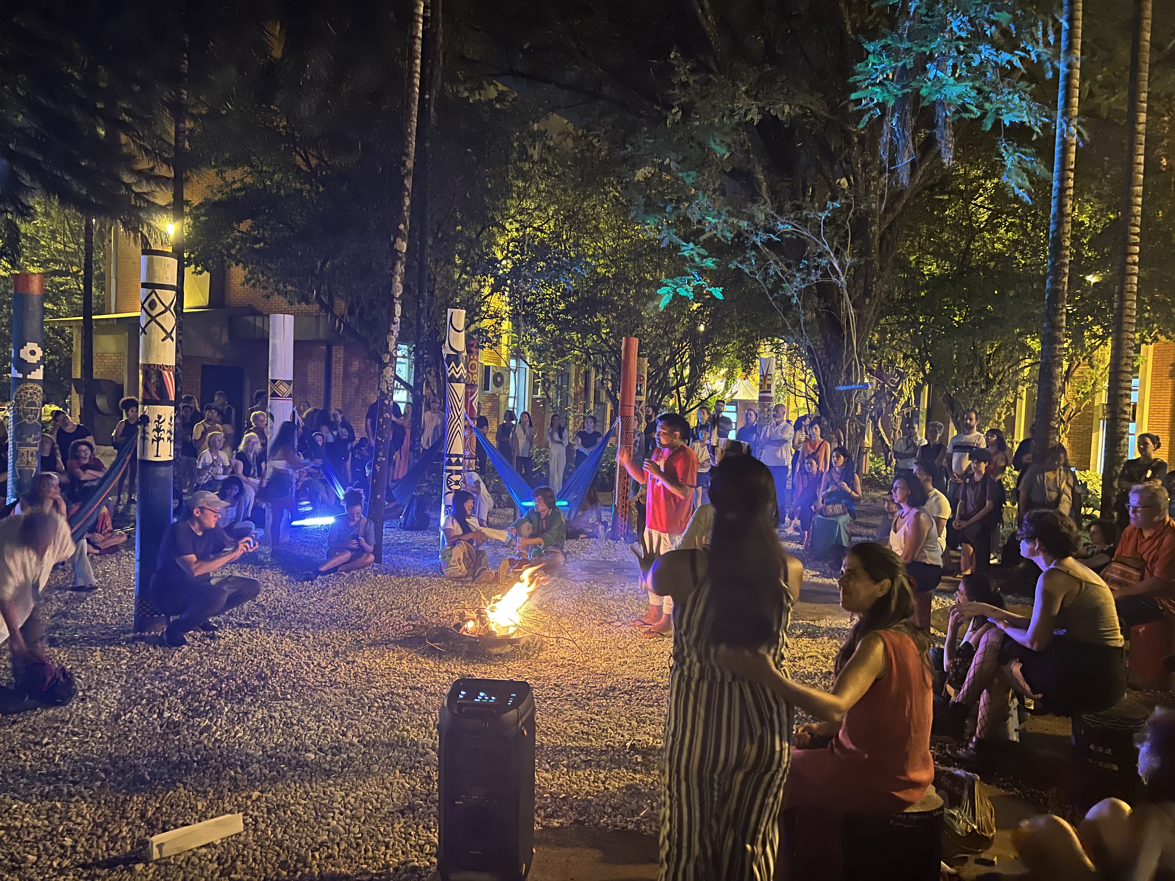 Fotografia do Jardim dos Saberes Ancestrais, à noite. Local ao ar livre, com chão de pedras brancas ao redor de círculos, mastros de madeira e bancos de madeira com grafismo indígenas coloridos. Pessoas sentadas no chão e em redes de tecido colorido. Ao centro, uma fogueira acesa. No fundo, paredes de tijolinhos, com janelas acesas.