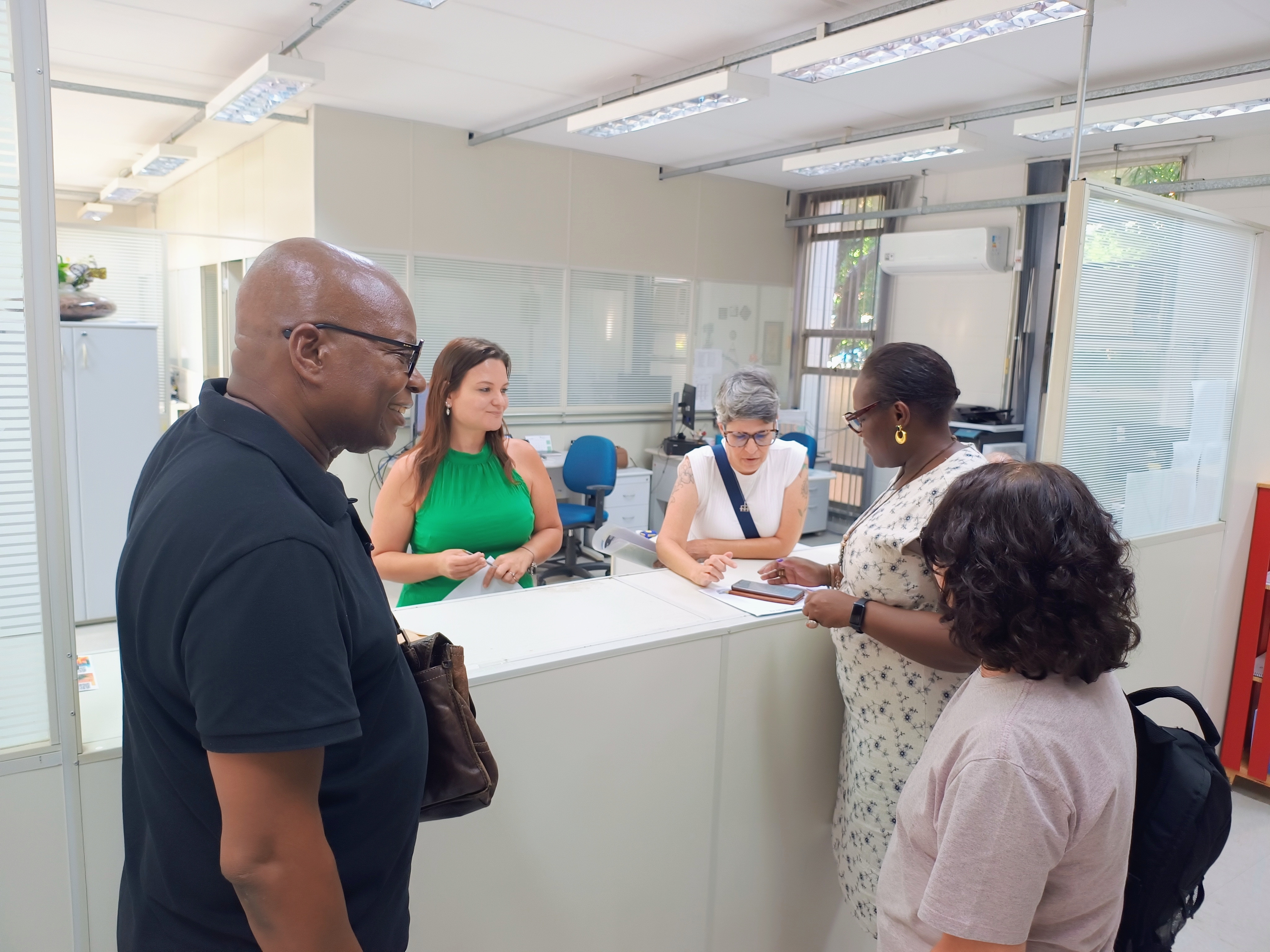 Fotografia do balcão de atendimento da Secretaria de Pós-Graduação. À direita, José Catiano, homem negro de óculo e camisa azul, sorrindo para as outras pessoas. Atrás do balcão duas mulheres atendem a diretora Debora Jeffrey, mulher negra com vestido branco florido. Ao seu lado, outra funcionária está de costas.