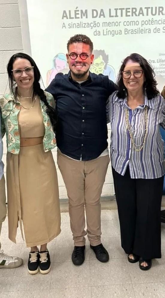 Fotografia do Prof. Dr. Guilherme Nichols, ao lado das orientadoras, em um auditório, com uma projeção de imagens na parede branca, onde lê-se "Além da Literatura Surda". Ele é um homem de pele bege média, com cabelos crespos escuros curtos, barba, óculos de grau redondo, camisa preta, calça bege.