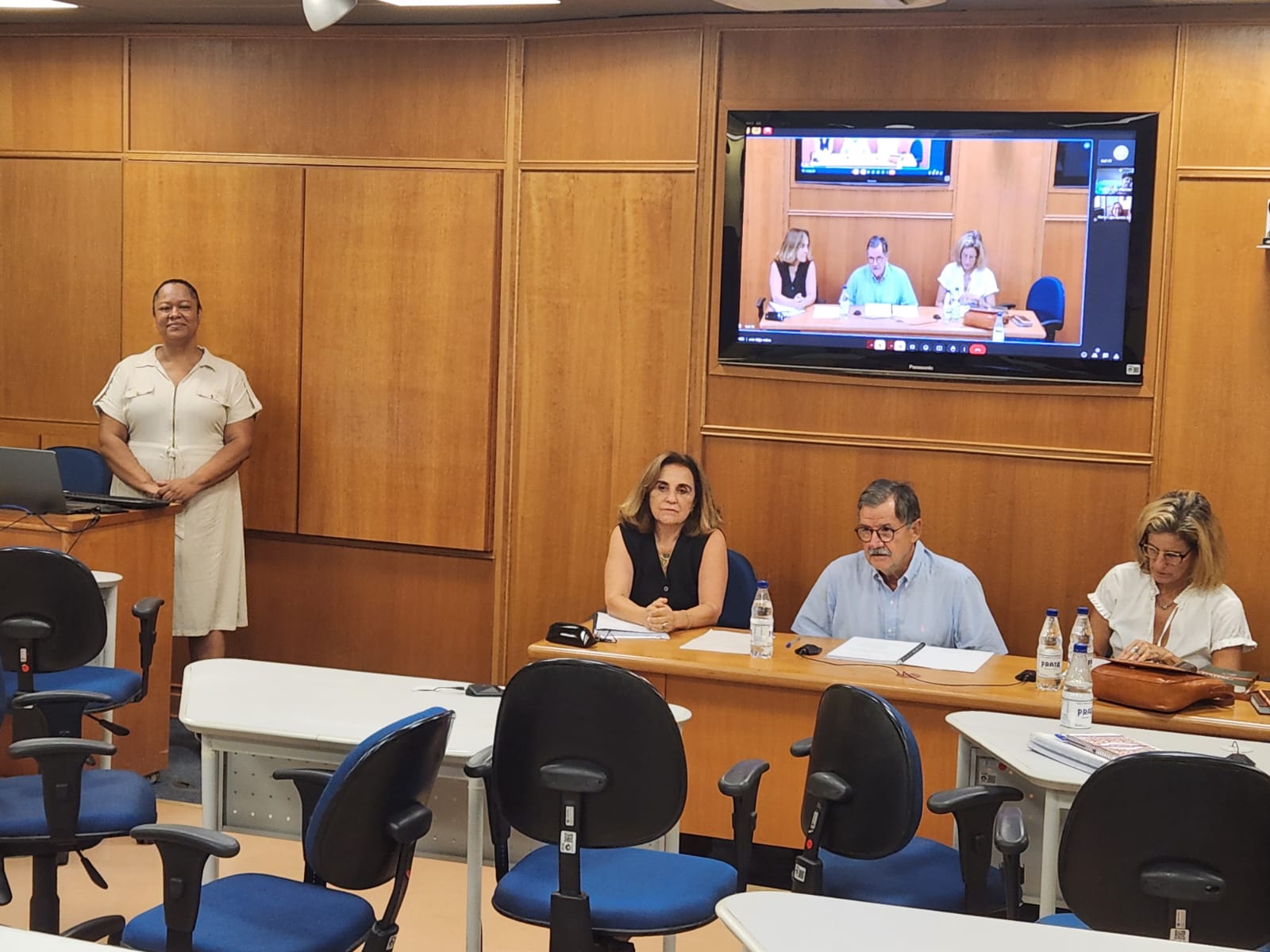 Fotografia de uma sala de conferência. Do lado esquerdo, uma mulher negra, de vestido bege, está em pé atrás de uma tribuna de madeira. Pessoas sentadas em uma mesa à frente do público fazem anotações. Ao fundo, parede de madeira, com monitor transmitindo a conferência em tempo real.
