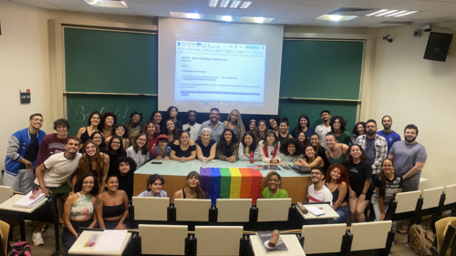 Uma grande quantidade de estudantes posa em uma sala de aula. Afrente da mesa principal há uma bandeira com faixas nas cores do arco-íris.