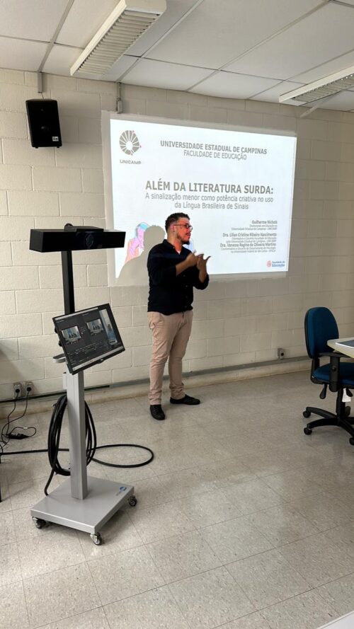 A imagem mostra um homem apresentando um trabalho acadêmico em uma sala de aula da Faculdade de Educação da Unicamp. Ele está em pé diante de um projetor, comunicando-se em Língua Brasileira de Sinais (Libras). No slide projetado está o título da apresentação: "ALÉM DA LITERATURA SURDA: A sinalização menor como potência criativa no uso da Língua Brasileira de Sinais", e o nome do apresentador, Guilherme Nichols, junto com os nomes das orientadoras e coorientadora. Ao lado do apresentador, há um equipamento de gravação ou transmissão com tela e câmera. A sala é clara, com paredes brancas e móveis simples, destacando o ambiente acadêmico e acessível.