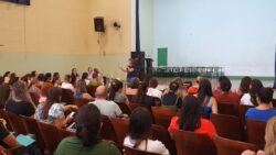 Uma imagem de um auditório lotado, com pessoas sentadas em cadeiras, enquanto uma palestrante se dirige ao grupo. O auditório é amplo, com inúmeras cadeiras dispostas em fileiras para acomodar o público. As cadeiras são de madeira escura, e o piso é claro. O público, composto por um grande grupo de pessoas, está sentado em silêncio, prestando atenção à palestra da palestrante, que está posicionada no palco, no centro da imagem. Ela está falando para o grupo e gesticulando. A composição da imagem é focada no público e na palestrante no palco, com o fundo levemente desfocado, criando um contraste claro entre a figura principal e o resto da cena. A iluminação é uniforme e natural, sem sombras marcantes. O foco da imagem é o grupo de pessoas, suas expressões e comportamentos. A maioria das pessoas está vestida com roupas casuais, típicas de um evento educacional. A palestrante está usando roupas mais formais. O ambiente é tranquilo e sério, com foco na escuta e no aprendizado do público. A imagem mostra um estilo fotográfico de rotina diária, sem detalhes específicos em cores ou estilos artísticos. Não há elementos que destaquem um estilo em especial na composição. O foco está na atmosfera do momento, e na ação central de uma mulher falando para um grupo. O ambiente é um auditório, possivelmente em uma escola ou centro comunitário. As paredes são de cor bege/creme, e o teto é de cor clara. Há uma área vazia no fundo, onde provavelmente está um palco ou espaço para apresentações. A iluminação é natural, sugerindo um dia ensolarado. A atmosfera é formal e atenciosa, adequada para um evento de educação ou treinamento.