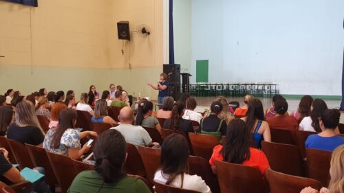 Uma imagem de um auditório lotado, com pessoas sentadas em cadeiras, enquanto uma palestrante se dirige ao grupo. O auditório é amplo, com inúmeras cadeiras dispostas em fileiras para acomodar o público. As cadeiras são de madeira escura, e o piso é claro. O público, composto por um grande grupo de pessoas, está sentado em silêncio, prestando atenção à palestra da palestrante, que está posicionada no palco, no centro da imagem. Ela está falando para o grupo e gesticulando. A composição da imagem é focada no público e na palestrante no palco, com o fundo levemente desfocado, criando um contraste claro entre a figura principal e o resto da cena. A iluminação é uniforme e natural, sem sombras marcantes. O foco da imagem é o grupo de pessoas, suas expressões e comportamentos. A maioria das pessoas está vestida com roupas casuais, típicas de um evento educacional. A palestrante está usando roupas mais formais. O ambiente é tranquilo e sério, com foco na escuta e no aprendizado do público. A imagem mostra um estilo fotográfico de rotina diária, sem detalhes específicos em cores ou estilos artísticos. Não há elementos que destaquem um estilo em especial na composição. O foco está na atmosfera do momento, e na ação central de uma mulher falando para um grupo. O ambiente é um auditório, possivelmente em uma escola ou centro comunitário. As paredes são de cor bege/creme, e o teto é de cor clara. Há uma área vazia no fundo, onde provavelmente está um palco ou espaço para apresentações. A iluminação é natural, sugerindo um dia ensolarado. A atmosfera é formal e atenciosa, adequada para um evento de educação ou treinamento.