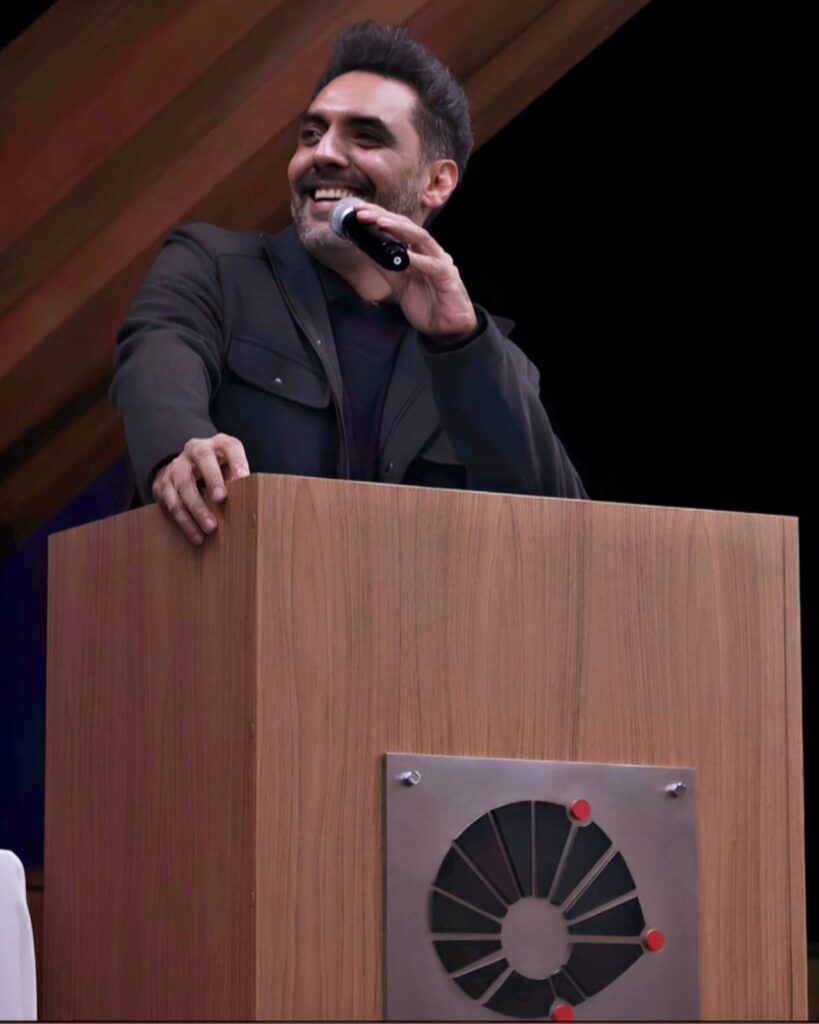 Um homem de pele média, com cabelos escuros e um sorriso amigável está em um palco, atrás de uma tribuna. Ele usa uma jaqueta escura e está falando ao microfone.  A tribuna é de madeira clara, com um painel metálico em formato circular no centro da mesa, contendo um padrão geométrico escuro, formando a logomarca da Unicamp. A iluminação e o fundo sugerem um ambiente interno formal de um auditório.