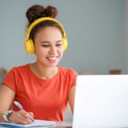 Uma moça sorridente usando fones de ouvido amarelos, camiseta laranja e cabelo preso em coque, sentada à mesa enquanto assiste a algo no notebook e escreve em um caderno.
