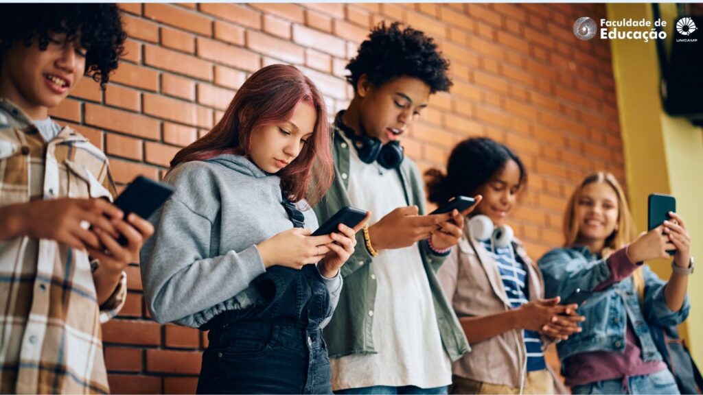 Uso de celulares nas escolas: Estudantes encostados numa parede, lado a lado, com a cabeça baixa utilizando celulares.