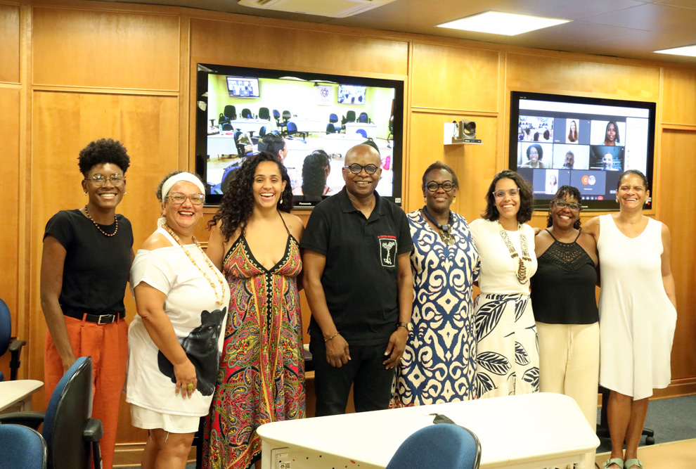 Na fotografia, há um grupo de pessoas, em uma aula aberta. Várias mulheres e um homem negro estão sorrindo e posam em pé para a foto, vestidos de forma casual. Há uma grande tela de televisão na parede atrás deles, com várias janelas mostrando diferentes atividades, paredes de madeira.