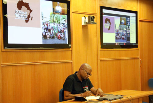 A imagem mostra um homem negro sentado em uma mesa, escrevendo em um caderno durante uma videoconferência. Ele veste uma camiseta preta com um logotipo no peito. Ele é o vice-reitor da Universidade Pedagógica de Maputo, José Castiano, que ministrou seminário na FE sobre a importância da aproximação entre Brasil e África. Atrás dele, há duas grandes telas na parede exibindo a mesma apresentação: o logotipo "Cor da Baixada" com o contorno do continente africano e uma foto de grupo de pessoas posando ao ar livre. O ambiente é uma sala com paredes de madeira clara, equipada para reuniões híbridas (presenciais e online).