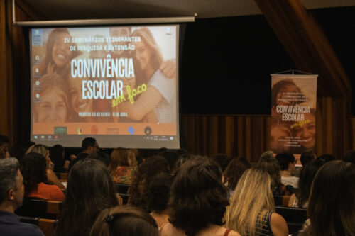 Fotografia de um auditório cheio de pessoas sentadas, assistindo a uma apresentação. A maioria dos presentes está de costas para a câmera, olhando para uma grande tela de projeção à frente. Na tela, aparece o slide de abertura de um evento chamado "IV Seminários Itinerantes de Pesquisa e Extensão – Convivência Escolar em Foco", com o subtítulo "Primeiro dia do evento – 01 de abril". A imagem de fundo do slide mostra jovens sorrindo, abraçados. Ao lado direito do auditório, há um banner vertical com o mesmo visual do slide, repetindo o nome do evento. O ambiente é fechado, com paredes de madeira e iluminação suave. O público é majoritariamente adulto e aparenta estar atento à programação.
