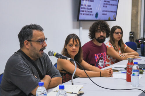 Quatro pessoas estão sentadas lado a lado em uma mesa clara, aparentemente participando de uma discussão ou apresentação. À esquerda, um homem de pele clara, com barba escura e óculos, fala ao microfone com os braços cruzados. Ao seu lado, uma mulher de pele clara e cabelos castanhos olha para ele com os braços apoiados na mesa. Próximo a ela, um homem de pele clara com cabelos cacheados escuros, barba e óculos veste uma camiseta bordô com uma estampa. A quarta pessoa, à direita, é uma mulher de pele clara com cabelos castanhos e óculos, vestindo uma blusa clara com uma estampa colorida e os braços cruzados. Sobre a mesa, há garrafas de água, cadernos e outros materiais. Ao fundo, uma parede branca serve de apoio para uma tela de televisão que exibe texto e imagens.
