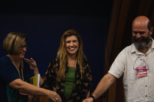 A imagem mostra três pessoas adultas, duas mulheres e um homem, sorrindo enquanto interagem entre si. A mulher ao centro está de frente para a câmera, com expressão alegre. Ela tem pele clara, cabelos loiros compridos e ondulados, e veste um vestido verde com um casaco preto estampado com flores vermelhas e bege. À esquerda, uma mulher de cabelos loiros curtos e óculos segura alguns cadernos e sorri olhando para o grupo. À direita, um homem com barba grisalha, também sorridente, veste uma camisa clara e tem um crachá rosa pendurado no pescoço com o nome “Guilherme”. As três pessoas parecem estar em um momento descontraído e de confraternização, possivelmente após uma palestra ou atividade, em um ambiente interno com fundo escuro e detalhes em madeira.
