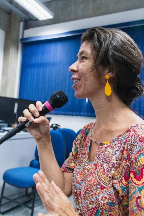 Uma mulher de pele clara com cabelos curtos e castanhos está em perfil, olhando para a esquerda e falando em um microfone preto com detalhes rosa. Ela veste uma blusa estampada com padrões coloridos em tons de vermelho, azul, amarelo e branco, e usa brincos amarelos em formato de gota. Suas mãos estão gesticulando enquanto ela fala. Ao fundo, parcialmente visíveis, há persianas azuis em uma janela e uma cadeira azul.