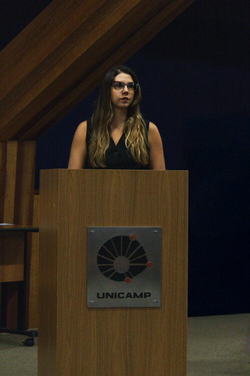 A imagem mostra uma mulher em pé atrás de um púlpito de madeira, no qual há uma placa metálica com o logotipo da UNICAMP (Universidade Estadual de Campinas) — um círculo segmentado com três pontos vermelhos e o nome da universidade abaixo. A mulher é jovem, de pele clara e cabelos longos castanhos com mechas claras. Ela usa óculos de armação marrom, blusa preta sem mangas e um colar discreto. A expressão no rosto é séria ou atenta, e ela olha para o lado direito da imagem, como se estivesse ouvindo ou aguardando algo. O fundo é escuro, com detalhes em madeira inclinada, típicos da arquitetura interna de auditórios.