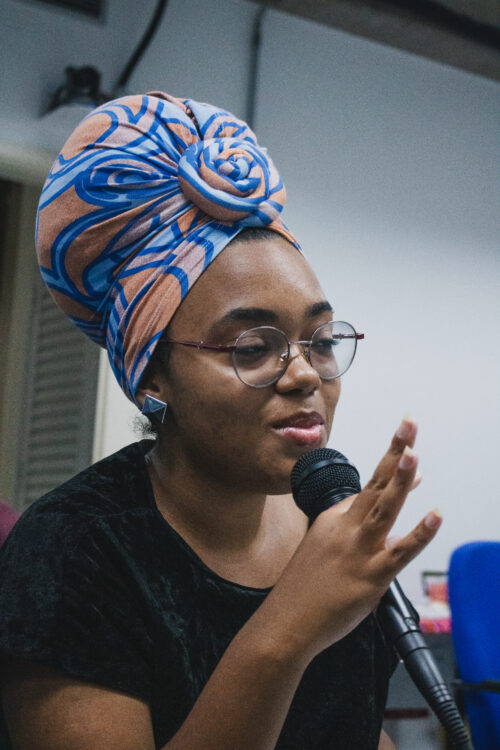 Uma mulher jovem e negra está em close-up, olhando levemente para a direita e falando em um microfone preto. Ela usa um turbante volumoso com estampas em tons de azul e laranja, além de óculos de armação fina e brincos geométricos prateados. Sua mão direita está próxima ao microfone, com os dedos levemente afastados, como se estivesse gesticulando enquanto fala. Ao fundo, um ambiente interno com cores neutras e partes de uma cadeira azul são visíveis.