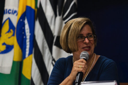A imagem mostra uma mulher de pele clara, cabelos lisos, loiros e curtos, com óculos de grau de armação escura, sorrindo enquanto fala ao microfone. Ela veste uma blusa azul escura, um colar e um anel dourado. A mulher está posicionada em frente a um conjunto de bandeiras — visíveis estão as bandeiras do Brasil, do estado de São Paulo, do município de Campinas e da Unicamp. O fundo é escuro e o foco está principalmente na mulher e nas bandeiras logo atrás dela.