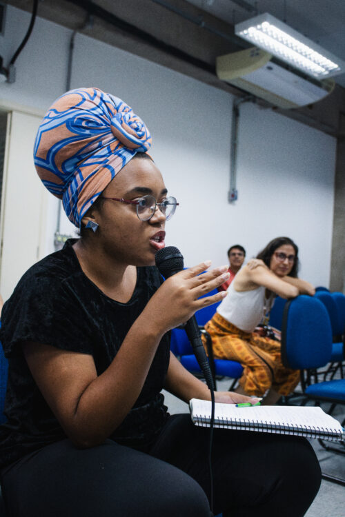 Uma pessoa negra está no centro da imagem, falando em um microfone preto. Ela usa um turbante volumoso com padrões em azul e laranja, óculos de armação fina e brincos geométricos prateados. Sua mão direita está próxima ao microfone, gesticulando enquanto fala. Ela está sentada em uma cadeira azul, com um caderno aberto sobre suas pernas. Ao fundo, desfocadas, estão outras pessoas sentadas em cadeiras azuis em um ambiente interno com paredes claras e iluminação artificial no teto.