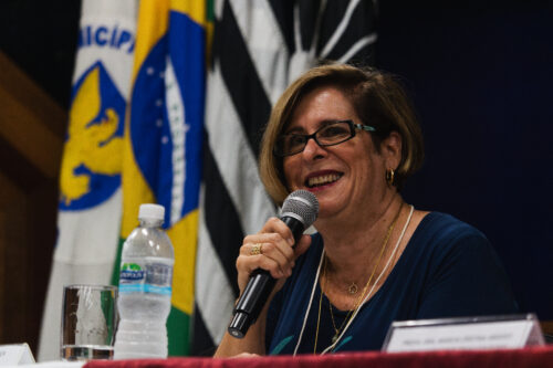 A imagem mostra a mesma mulher da imagem anterior. Ela está sorrindo enquanto segura um microfone perto da boca. A mulher tem pele clara, cabelo loiro curto com luzes, usa óculos de grau escuros, brincos dourados e colares. Está vestindo uma blusa azul escura. À sua frente, sobre a mesa, há uma garrafa de água, um copo e uma plaquinha parcialmente visível. Ao fundo, aparecem as mesmas bandeiras: do Brasil, do estado de São Paulo, do município de Campinas e da Unicamp
