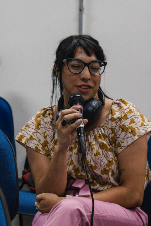 Uma pessoa de pele clara com cabelos escuros e franja está sentada, segurando um microfone preto com detalhes rosa e falando. Ela usa óculos de armação preta, um piercing no nariz, brincos pequenos e um anel escuro em um dos dedos. Um fone de ouvido azul escuro está pendurado em seu pescoço. Ela veste uma blusa de fundo claro com estampas florais em tons de amarelo e marrom, e calças rosa claro. Ao fundo, parcialmente visíveis, há paredes claras e cadeiras azuis. A iluminação parece suave, vinda de cima.