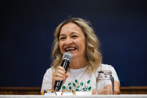 A imagem mostra uma mulher sorridente, com cabelos claros e ondulados, na altura dos ombros. Ela está segurando um microfone com a mão direita e parece estar falando com entusiasmo, mostrando os dentes. Ela veste uma camiseta branca com um desenho de folhas verdes estilizadas na frente. No pescoço, ela usa um colar delicado com um pingente redondo. À frente dela, sobre uma mesa, com toalha branca, podemos ver um par de óculos com armação laranja e branca e um copo de vidro transparente, virado para baixo. O fundo da imagem é um azul escuro uniforme. O ambiente é o palco do Centro de Convenções da Unicamp.