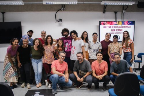 Um grupo de dezesseis pessoas está reunido para uma foto em um ambiente interno com iluminação artificial no teto. A maioria está em pé, lado a lado, enquanto alguns estão agachados na frente. As pessoas apresentam diversas características físicas e vestimentas, com uma variedade de tons de pele, tipos de cabelo e estilos de roupa, incluindo camisetas, calças jeans e saias estampadas. Ao fundo, há uma parede clara com uma tela de televisão mostrando uma apresentação com texto e imagens coloridas, além de outros elementos como um quadro branco e equipamentos eletrônicos. Cadeiras azuis estão dispostas na sala. A atmosfera geral sugere um evento informal ou uma reunião de grupo.