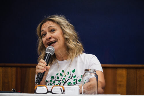 A imagem apresenta uma mulher de cabelos loiros e ondulados, na altura dos ombros, que sorri e olha para a esquerda enquanto fala ou canta com entusiasmo em um microfone preto que segura com a mão direita. Ela veste uma camiseta branca com o desenho de uma árvore verde estilizada e um colar com um pingente claro. À sua frente, sobre uma mesa branca, há óculos com armação preta e branca e um copo de vidro virado para baixo. O fundo é composto por uma parede azul escura acima e painéis de madeira marrom abaixo.