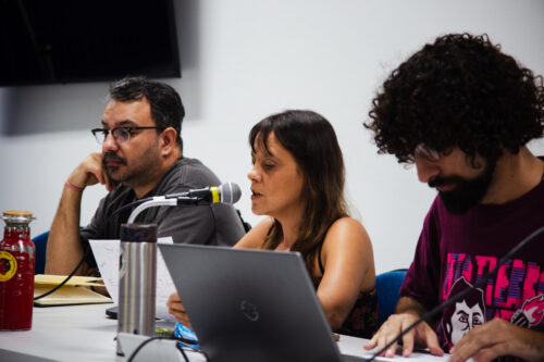 Três pessoas estão sentadas lado a lado em uma mesa clara, engajadas em uma discussão. À esquerda, um homem de pele clara com barba escura e óculos apoia o rosto na mão, parecendo ouvir atentamente. No centro, uma mulher de pele clara e cabelos castanhos fala ao microfone posicionado à sua frente, com um laptop aberto em sua frente. À direita, um homem de pele clara com cabelos cacheados escuros e barba está inclinado sobre a mesa, olhando para papéis. Sobre a mesa, além dos microfones e do laptop, há garrafas, papéis e outros objetos. O fundo é uma parede branca com um objeto escuro na parte superior esquerda. A iluminação parece vir de cima.