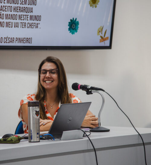 Uma mulher de pele clara com cabelos castanhos e óculos está sentada a uma mesa clara, sorrindo com os olhos fechados. À sua frente, há um laptop cinza aberto e um microfone preto com detalhes rosa. Uma garrafa térmica prateada com adesivos está à sua esquerda. Ao fundo, uma parede clara com uma tela de televisão parcialmente visível exibe texto e imagens coloridas. A iluminação parece suave e difusa.