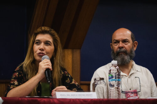 A imagem apresenta duas pessoas sentadas lado a lado atrás de uma mesa coberta por um tecido vermelho escuro. À esquerda, uma mulher de pele clara com cabelos longos e ondulados em tons de castanho claro e loiro está falando ao microfone, que segura com a mão direita. Ela veste uma blusa escura com um padrão floral em tons de vermelho e verde. À direita dela, um homem com barba e espessa olha para a esquerda. Ele veste uma camisa de cor clara e tem óculos pendurados por um cordão no pescoço. Sobre a mesa, à frente da mulher, há uma placa com o nome "PROFA. DRA. MARCIA CRISTINA ARGENTI". Entre os dois, há uma garrafa de água de plástico transparente e um pote de vidro.