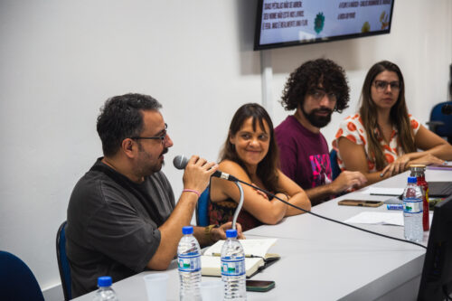Quatro pessoas estão sentadas lado a lado em uma mesa clara, participando de uma conversa ou apresentação. À esquerda, um homem de pele clara, com barba escura e óculos, fala ao microfone com a mão direita levantada. Ao seu lado, uma mulher de pele clara e cabelos castanhos sorri com os braços cruzados sobre a mesa. Próximo a ela, um homem de pele clara com cabelos cacheados escuros, barba e óculos olha para a esquerda. A quarta pessoa, à direita, é uma mulher de pele clara com cabelos castanhos e óculos, vestindo uma blusa clara com estampa de frutas e os braços apoiados na mesa. Sobre a mesa, há garrafas de água, papéis e outros objetos. Ao fundo, uma parede branca serve de apoio para uma tela de televisão que exibe texto e imagens. A iluminação ambiente é clara.