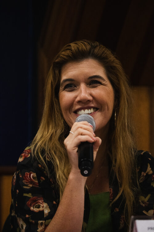 A imagem mostra uma mulher sorridente com cabelos longos e ondulados em tons de castanho claro e loiro. Ela está segurando um microfone com a mão direita, bem perto da boca, como se estivesse falando ou cantando com alegria, mostrando os dentes. Ela veste uma blusa escura com um padrão de flores grandes em tons de vermelho e verde. Podemos ver um pouco de uma outra peça de roupa verde por baixo da blusa. Ela usa brincos de argola dourados e um anel prateado na mão que segura o microfone. O fundo da imagem é parcialmente visível e mostra painéis de madeira na parte superior e uma área mais escura abaixo.