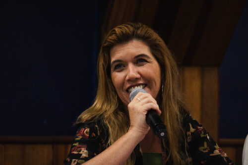 A imagem mostra a mesma da imagem anterior. Uma mulher sorridente com cabelos longos e ondulados em tons de castanho claro e loiro. Ela segura um microfone com a mão direita, posicionado perto de sua boca, como se estivesse falando ou cantando com entusiasmo, exibindo seus dentes. Ela veste uma blusa escura adornada com um padrão de flores grandes em tons de vermelho e verde. É possível notar uma peça de roupa verde por baixo da blusa. A mulher utiliza brincos de argola dourados e um anel prateado na mão que segura o microfone.