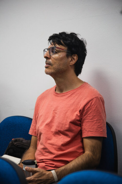 Um homem de pele clara com cabelos escuros e óculos está sentado em uma cadeira azul, olhando para a esquerda em perfil. Ele veste uma camiseta laranja e parece estar ouvindo ou observando algo fora do campo de visão. Suas mãos estão juntas no colo. Ao fundo, uma parede branca lisa é visível. A iluminação parece suave e indireta.