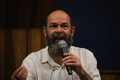 A imagem apresenta um homem de pele clara, com barba grisalha e espessa e bigode. Ele está segurando um microfone com a mão direita, posicionado perto da boca, como se estivesse falando. Sua mão esquerda está levemente levantada e aberta, gesticulando enquanto fala. Ele veste uma camisa de cor clara com botões. Um cordão de óculos preto está visível ao redor de seu pescoço. Ele também usa um anel escuro no dedo anelar da mão esquerda. O fundo da imagem é dividido horizontalmente, com painéis de madeira em tons de marrom na parte superior e uma área azul escura e uniforme abaixo.