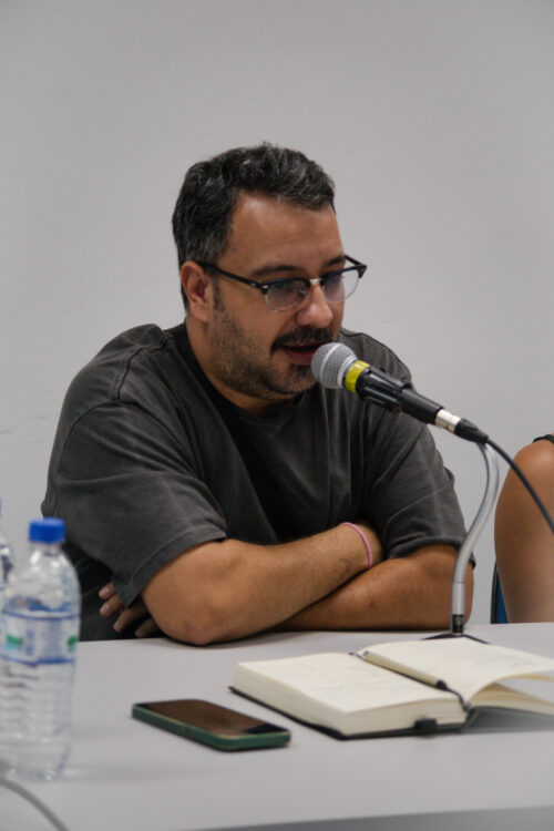 Um homem de pele clara, com barba escura e óculos, está sentado a uma mesa clara, falando em um microfone preto com detalhes prateados. Seus braços estão cruzados sobre a mesa, onde também se encontram uma garrafa de água com tampa azul, um smartphone preto e um caderno aberto com páginas brancas. Ao fundo, uma parede branca lisa é visível. A iluminação parece vir de cima, criando sombras suaves.