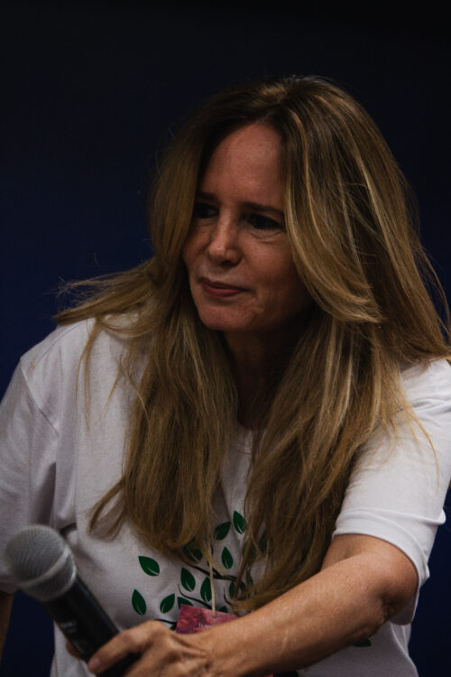 A imagem apresenta uma mulher de pele clara com longos cabelos castanhos claros e loiros, levemente ondulados, que caem sobre seus ombros. Ela parece estar olhando para algo à sua esquerda, com uma expressão calma e um leve sorriso. Ela veste uma camiseta branca com uma estampa de folhas verdes e um elemento rosa na parte inferior. Sua mão direita segura a parte inferior de um microfone preto, que está parcialmente visível na parte inferior esquerda da imagem. O fundo é um azul escuro e uniforme, o que destaca a mulher como o foco principal da fotografia.