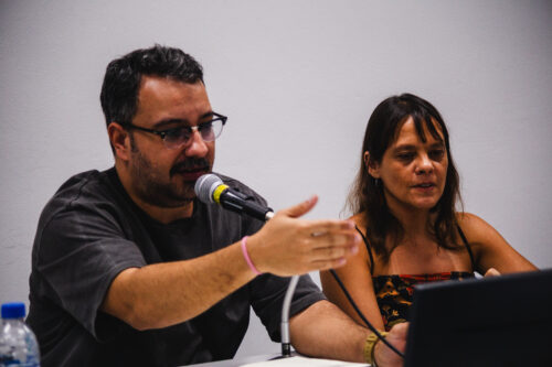 Dois indivíduos estão sentados lado a lado em uma mesa clara. À esquerda, um homem de pele clara com barba escura e óculos fala ao microfone, gesticulando com a mão direita aberta. Ele veste uma camiseta cinza escura e uma pulseira rosa no pulso esquerdo. À sua direita, uma mulher de pele clara com cabelos castanhos olha em sua direção, com parte de um laptop preto visível à sua frente. Ela veste uma blusa escura com detalhes coloridos. Uma garrafa de água com tampa azul está sobre a mesa à esquerda do homem. Ao fundo, uma parede branca lisa é visível. A iluminação parece vir de cima.