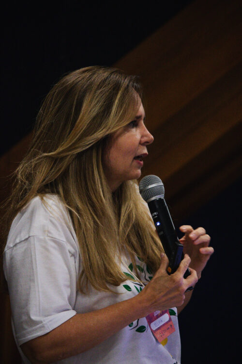 A imagem apresenta uma mulher de perfil, com longos cabelos castanhos claros e loiros que caem sobre seus ombros. Ela está falando ao microfone, que segura com ambas as mãos perto da boca. Sua expressão facial é de concentração enquanto fala. Ela veste uma camiseta branca com uma estampa de folhas verdes e um elemento rosa na parte inferior. Presa à sua camiseta, há um crachá com texto ilegível. O fundo da imagem é escuro na parte inferior e possui painéis de madeira em tons de marrom na parte superior.