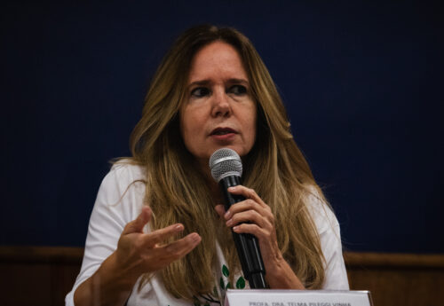A imagem apresenta uma mulher de pele clara com longos cabelos castanhos claros e loiros, que caem sobre seus ombros. Ela está falando ao microfone, que segura com a mão direita, enquanto gesticula com a mão esquerda aberta. Sua expressão facial é séria e focada enquanto fala. Ela veste uma blusa branca com mangas compridas. À sua frente, sobre uma superfície clara, há uma placa com o texto "PROFA. DRA. TELMA PILEGGI VINHA". O fundo da imagem é um azul escuro e uniforme. A iluminação parece vir da frente, iluminando o rosto e a parte superior do corpo da mulher.