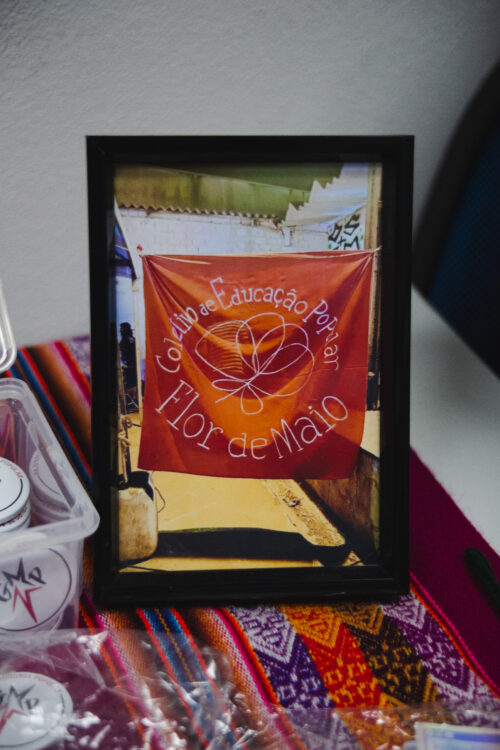 Uma fotografia emoldurada com uma moldura preta está sobre uma superfície colorida com padrões geométricos em tons de vermelho, laranja, azul e branco. A fotografia retrata um banner de cor laranja/avermelhada pendurado em um ambiente externo, com o texto bordado em branco "Coletivo de Educação Popular" em um semicírculo na parte superior e "Flor de Maio" em letras maiores na parte inferior, acompanhado de um desenho estilizado de uma flor branca. Ao redor do banner, é possível observar elementos do ambiente externo, como paredes e outros objetos não identificados. Em primeiro plano, parcialmente visíveis, há objetos diversos, incluindo um recipiente plástico transparente e embalagens. A iluminação parece natural, incidindo sobre a fotografia e a superfície onde ela está apoiada.