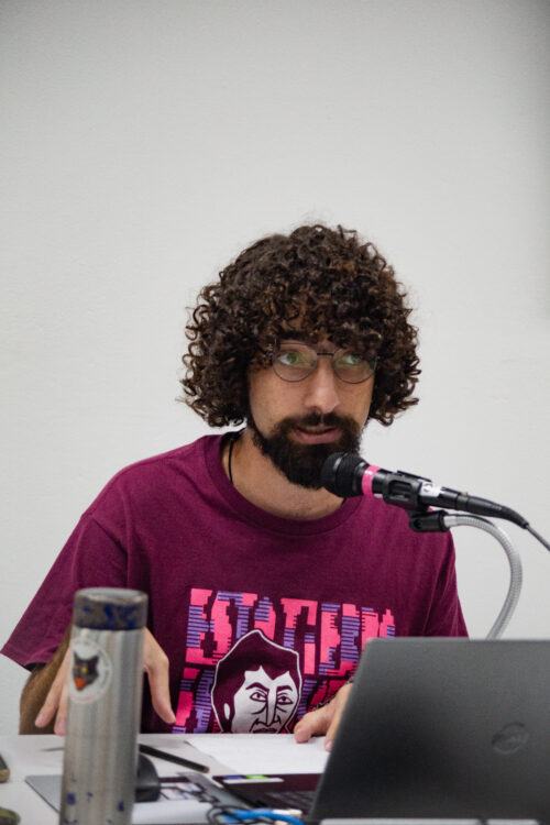 Um homem de pele clara com cabelos cacheados escuros, barba e óculos está posicionado em frente a um microfone preto com detalhes rosa, olhando levemente para a direita. Ele veste uma camiseta bordô com uma estampa colorida em tons de rosa e azul. À sua frente, parcialmente visíveis, há um laptop cinza aberto e uma garrafa térmica prateada com adesivos. O fundo é uma parede branca lisa. A iluminação parece vir de cima, focando no homem.