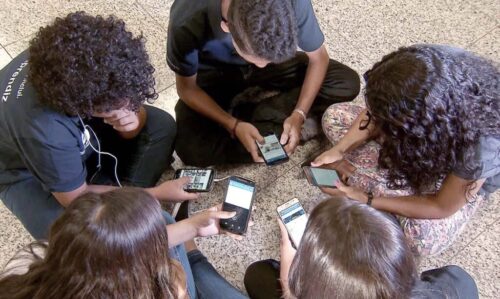 Seis jovens sentados em círculo no chão, todos concentrados em seus celulares.