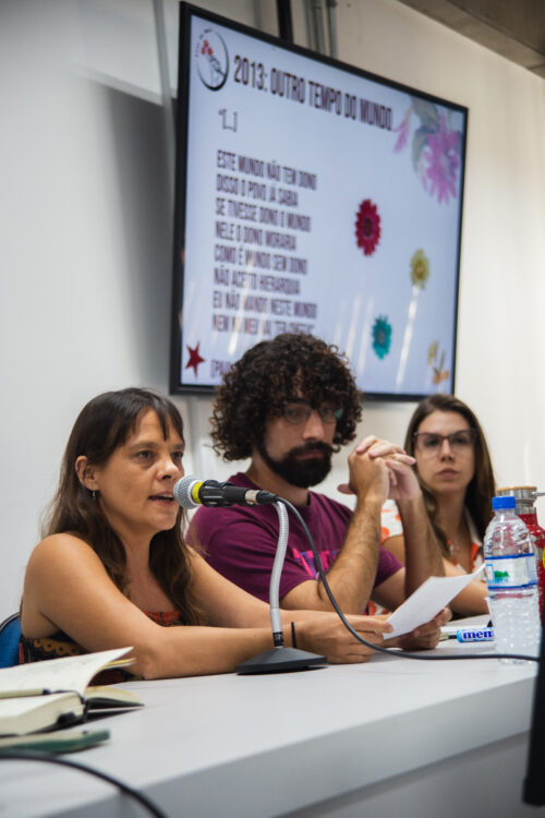 A imagem mostra três pessoas sentadas lado a lado em uma mesa de debate. À esquerda, uma mulher de pele clara, cabelos lisos e castanhos escuros, veste um vestido sem mangas estampado em tons terrosos. Ela está falando ao microfone e segurando um papel. No centro, há um homem de pele clara, cabelo cacheado e barba escura, usando óculos e uma camiseta vinho. Ele está com as mãos entrelaçadas e expressão atenta. À direita, uma mulher de pele clara, cabelo liso e castanho claro, veste óculos e camisa branca com detalhes laranja. À frente deles há garrafas, copos e um microfone. Ao fundo, há uma tela com a projeção do título “2013: OUTRO TEMPO DO MUNDO”, seguido de um trecho de poema atribuído a Paulo César Pinheiro. A tela tem também desenhos de flores coloridas.