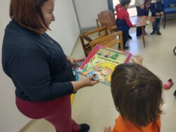Fotografia de uma mulher segurando o livro infantil “O Príncipe Sapo” e uma criança interagindo com o livro, na Biblioteca da Faculdade de Educação da UNICAMP. A criança é um menino de pele clara, tem cabelo castanho e liso e veste uma blusa laranja, e está apontando para a capa do livro. A mulher tem pele clara, cabelo castanho e liso e veste uma blusa preta e uma calça rosa, e está sorrindo. A capa do livro contém uma ilustração de uma moça loira sendo beijada na bochecha por um sapo. Ao fundo da fotografia, há crianças ao redor de uma mesa brincando com papéis. Fim da descrição.