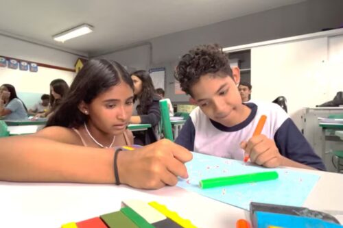 Ao lado de uma colega, Pedro Domingues desenha segurando um lápis de cor grosso, com diâmetro maior, na cor laranja.